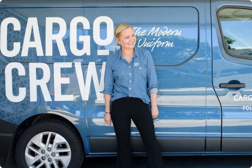smile blonde woman in blue shirt and black pants stands in front of 'Cargo Crew - the modern uniform' van