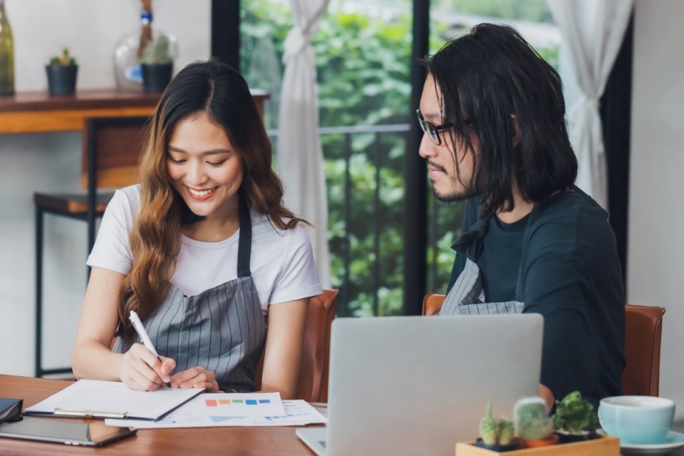 smiling woman marks off a checklist while sitting beside a man with an open laptop on a table in front of them